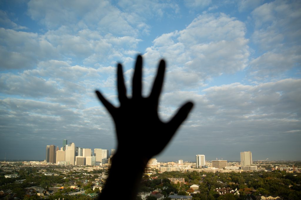 A hand is silhouetted reaching out against a window with a downtown skyline seen beyond.