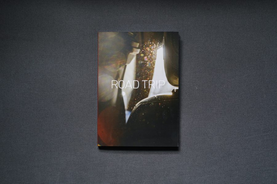 Photograph of the ROAD TRIP printed publication on a dark grey background. The cover shows a photograph of light hitting dust between the headrest of a front seat and the left side of a car interior, with lens flare. Text saying 'ROAD TRIP' in white font is in the centre of the image.