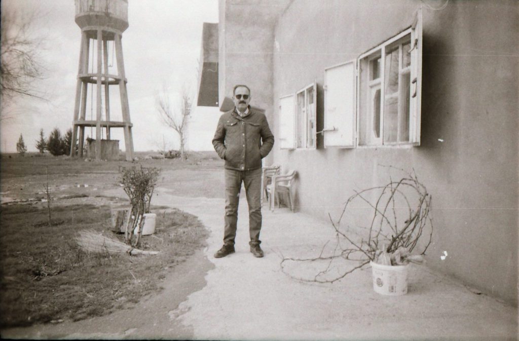 an elderly man stands on a path in front of a building, behind him is a large water tower