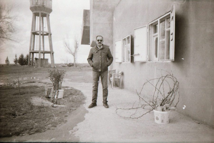 an elderly man stands on a path in front of a building, behind him is a large water tower