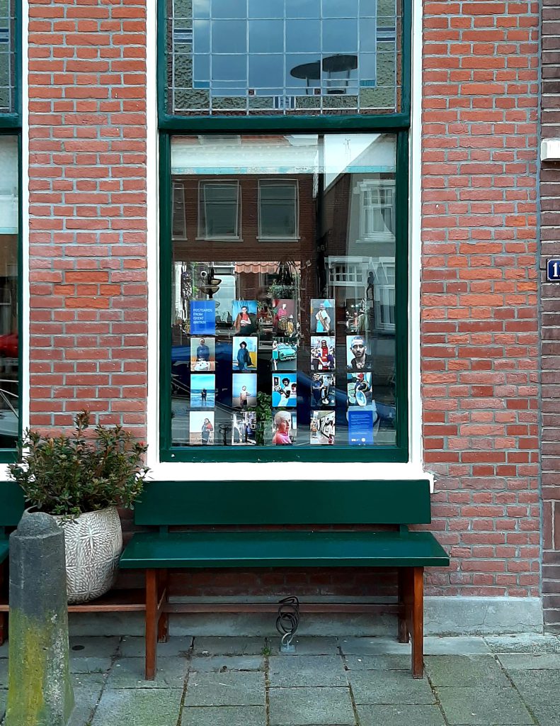 Photograph of a window displaying postcard-sized photographs, with brick wall either side, a green wooden bench in front and a potted plant to the left.
