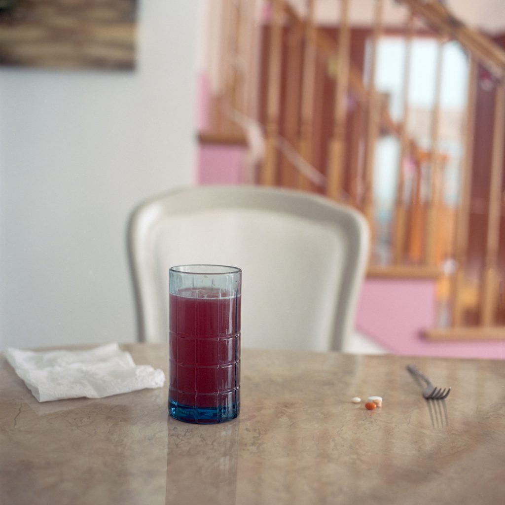Photograph of a blue glass with raspberry iced tea, a napkin, vitamins, and a fork set at a marbled table with a chair and a set of stairs behind it.
