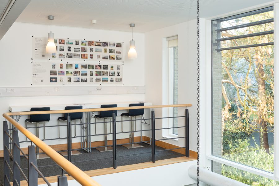 Installation photograph of 'Postcards from Europe' exhibition showing postcard-sized photographs printed on a long strip of newsprint taped to the wall, with lampshades suspended from the ceiling in front, stools below, a railing in front and a window showing plants outside to the right.