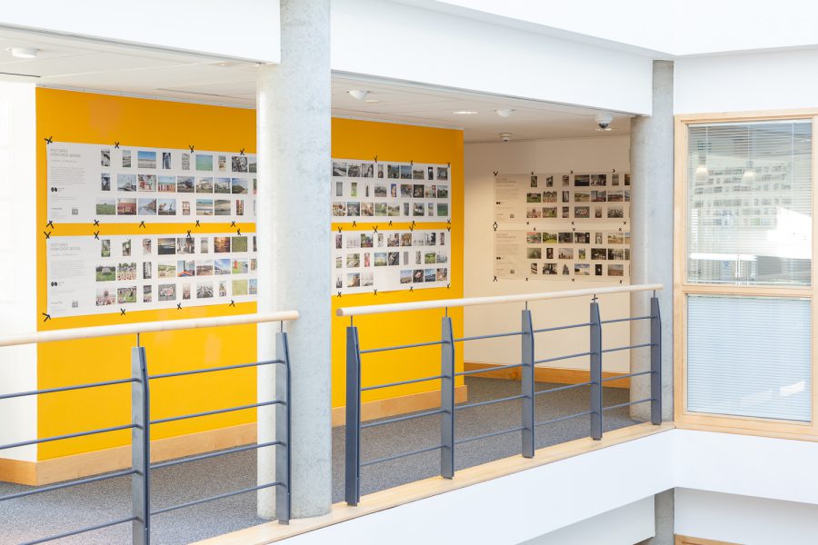 Installation photograph of 'Postcards from Europe' exhibition, showing postcard-sized photographs printed on long strips of newsprint, taped to a yellow wall and a white all further in the distance, with concrete pillars and railings in front.