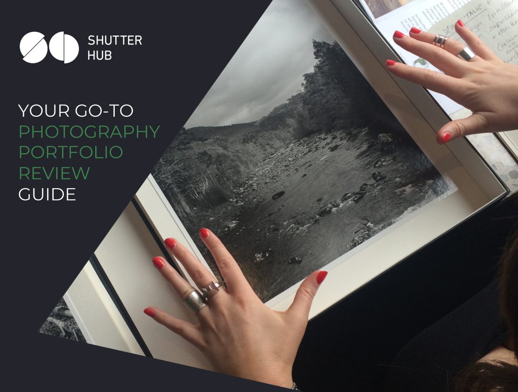 Graphic with the Shutter Hub logo top left, text reading 'YOUR GO-TO PHOTOGRAPHY PORTFOLIO REVIEW GUIDE' below in white and green text and a photograph to the right showing hands with red painted fingernails and silver rings above a printed photograph in a box, the black and white photograph appearing to show a river with rocks in it surrounded by trees and mountains.