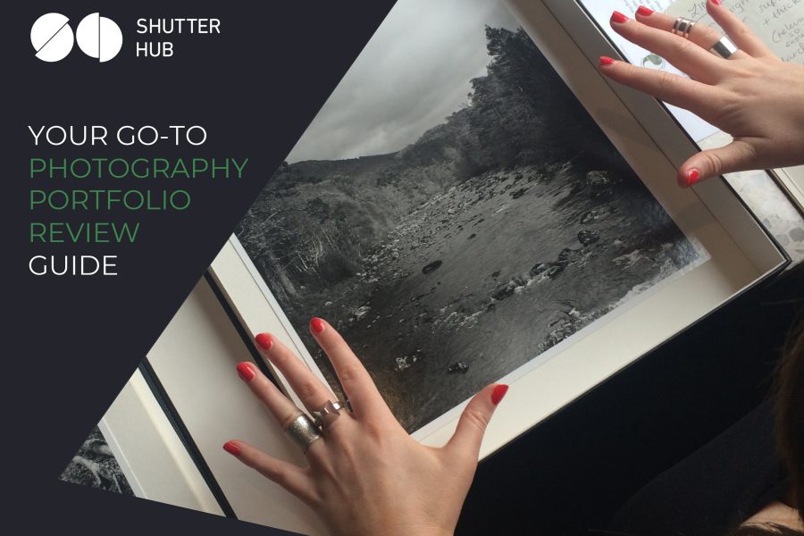 Graphic with the Shutter Hub logo top left, text reading 'YOUR GO-TO PHOTOGRAPHY PORTFOLIO REVIEW GUIDE' below in white and green text and a photograph to the right showing hands with red painted fingernails and silver rings above a printed photograph in a box, the black and white photograph appearing to show a river with rocks in it surrounded by trees and mountains.
