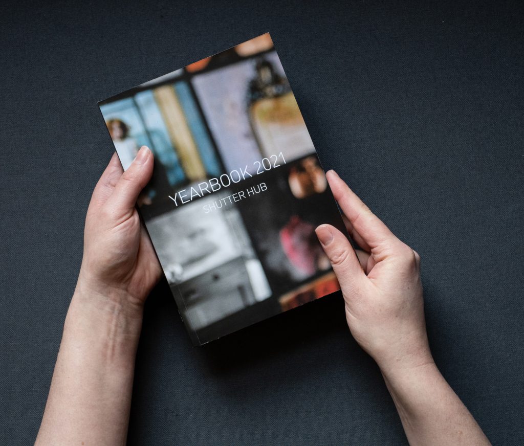 Photograph of hands holding a copy of the YEARBOOK printed publication, with the text 'YEARBOOK 2021', Shutter Hub' on the cover, against a dark grey fabric background
