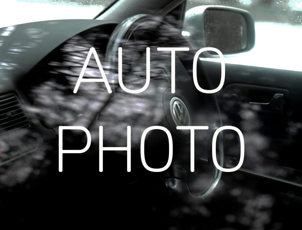 Graphic with the text 'AUTO PHOTO' in white in the centre, and in the background a photograph of a steering wheel of a Volkswagen Gold seen through the passenger side window, reflections of trees are visible on the glass.