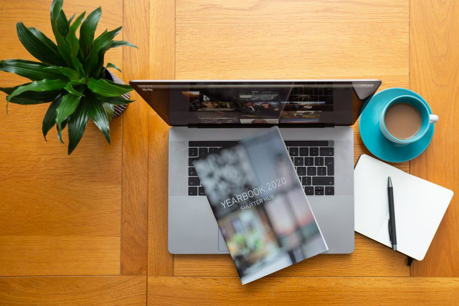 Looking down at a table with a pot plant to the left, laptop with YEARBOOK printed publication on top, a cup of coffee and a notebook with a pen to the right