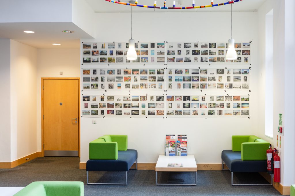 Installation photograph of 'Postcards from Europe' exhibition, showing postcard-sized photographs printed on long strips of newsprint, taped to the wall, with a lighting installation suspended from the ceiling above, green and blue sofas and a table below and a wooden door to the left.