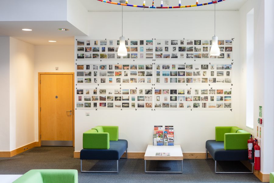 Installation photograph of 'Postcards from Europe' exhibition, showing postcard-sized photographs printed on long strips of newsprint, taped to the wall, with a lighting installation suspended from the ceiling above, green and blue sofas and a table below and a wooden door to the left.