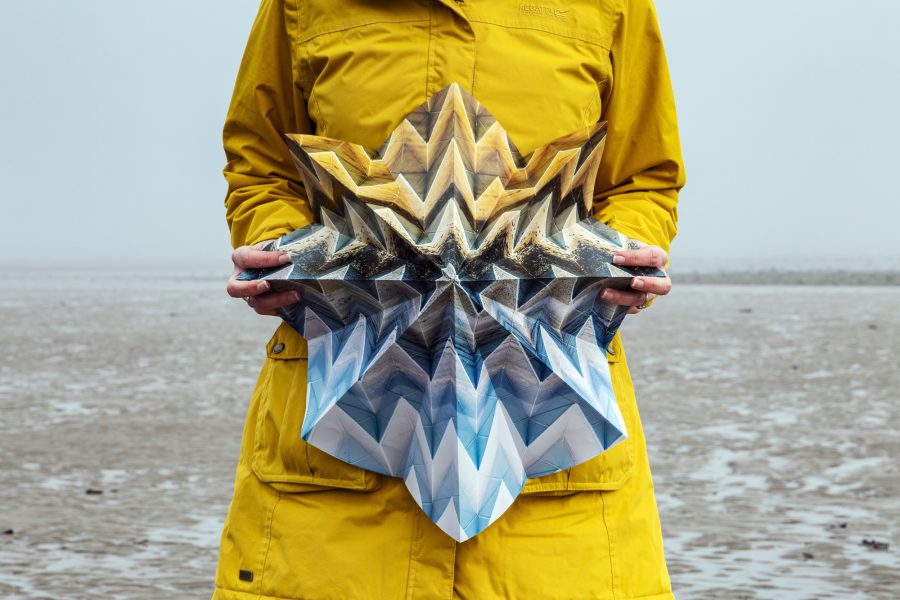 Image of a woman standing on a foggy beach and holding a large, intricately-folded, photograph that blends in with the landscape.