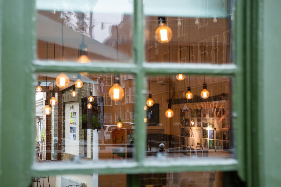 Photograph looking through a window into the Gallery Café at St. Margaret's House, showing an exhibition of photographs printed on newsprint, and light bulbs hanging from the ceiling