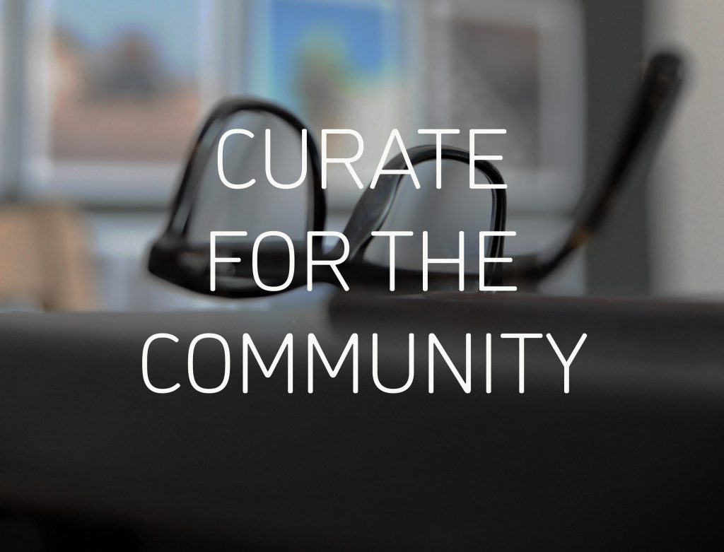 Graphic reading 'CURATE FOR THE COMMUNITY' in white text in the centre. Behind is a photograph of black framed glasses upside down on a dark surface, with framed pictures visible but out of focus in the background.