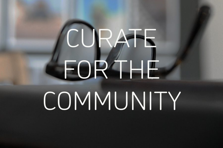 Graphic reading 'CURATE FOR THE COMMUNITY' in white text in the centre. Behind is a photograph of black framed glasses upside down on a dark surface, with framed pictures visible but out of focus in the background.